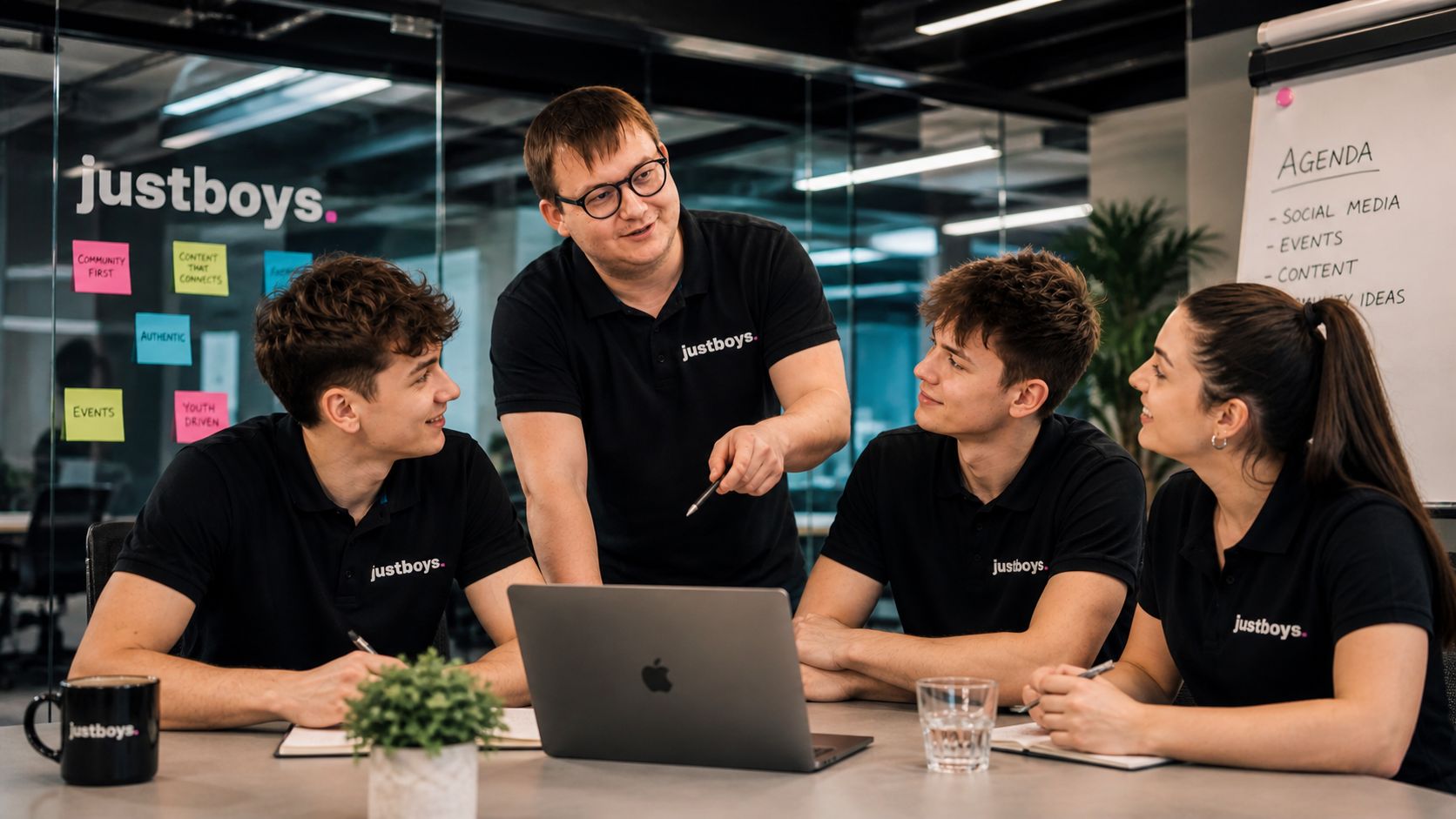 Vier Personen der justboys-Redaktion in schwarzen Polos am Konferenztisch - am Whiteboard im Hintergrund Themen wie Social Media, Events und Content.